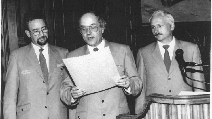 Eine Schwarz-Weiß-Aufnahme von drei Männern, vor der linken Person steht ein Mikrofon, der Mann in der Mitte hält ein großes Dokument, das er vorliest. (Quelle: THW) Eine Schwarz-Weiß-Aufnahme von drei Männern, vor der linken Person steht ein Mikrofon, der Mann in der Mitte hält ein großes Dokument, das er vorliest.