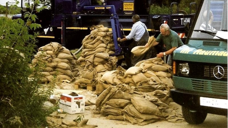 Im Vordergrund liegen aufeinander gestapelte Sandsäcke. Dahinter stehen zwei Personen, die Sandsäcke füllen und aufreihen. Im Hintergund steht ein blaues Fahrzeug und neben den aufgereihten Sandsäcken parkt ein grünes Fahrzeug. (Quelle: THW) Im Vordergrund liegen aufeinander gestapelte Sandsäcke. Dahinter stehen zwei Personen, die Sandsäcke füllen und aufreihen. Im Hintergund steht ein blaues Fahrzeug und neben den aufgereihten Sandsäcken parkt ein grünes Fahrzeug.