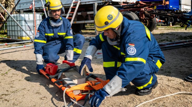 Zwei Personen mit blauer THW-Einsatzkleidung und gelben Helmen hocken vor einer orangen Trage. Auf der Trage liegt ein Gewicht aus Holz. Im Hintergrund stehen mehrere große Anhänger. (Quelle: THW/Max Mölkner) Zwei Personen mit blauer THW-Einsatzkleidung und gelben Helmen hocken vor einer orangen Trage. Auf der Trage liegt ein Gewicht aus Holz. Im Hintergrund stehen mehrere große Anhänger.
