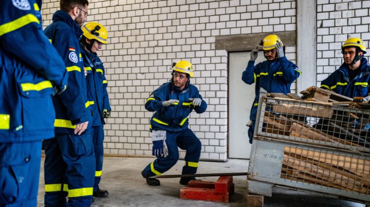 Mehrere Menschen in Einsatzkleidung stehen in einer Halle. Eine Gitterbox mit Holz steht mit einer Seite auf einem Stück Holz. Ein Mensch steht daneben und geht in die Knie. (Quelle: THW/Max Mölkner) Mehrere Menschen in Einsatzkleidung stehen in einer Halle. Eine Gitterbox mit Holz steht mit einer Seite auf einem Stück Holz. Ein Mensch steht daneben und geht in die Knie.