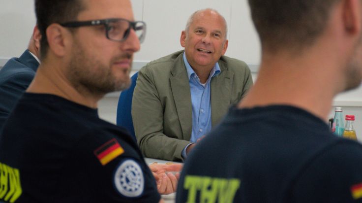 Ein Mensch sitzt an einem Tisch. Ihm gegenüber sitzen zwei weitere Menschen, die von hinten und angeschnitten zu sehen sind. (Quelle: THW/Alessandro Geßner) Ein Mensch sitzt an einem Tisch. Ihm gegenüber sitzen zwei weitere Menschen, die von hinten und angeschnitten zu sehen sind.