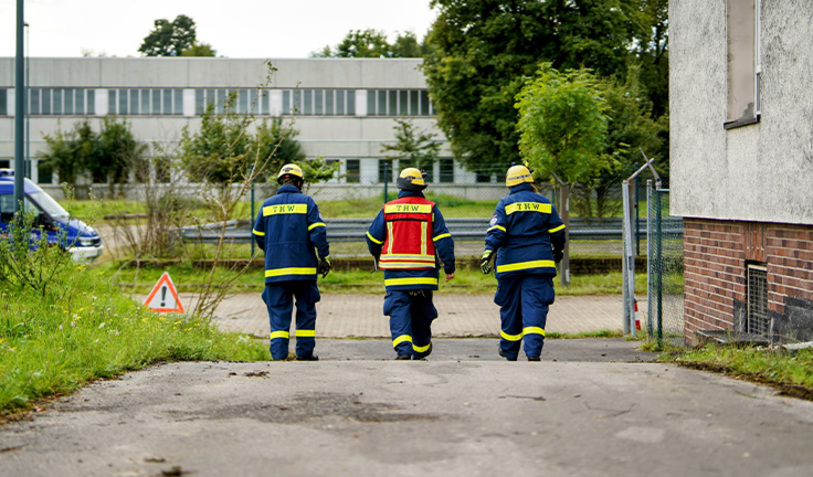 Drei THW-Kräfte in blauer Einsatzkleidung gehen eine Straße entlang. Alle drei tragen gelbe Helme. Die Person in der Mitte trägt eine rote Weste mit der Aufschrift "THW".