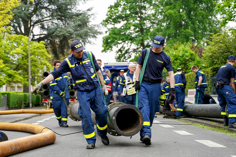 Im Vordergrund des Bildes gehen zwei THW-Kräfte in blauer Einsatzkleidung, die gemeinsam mit einer grünen Halterung ein Rohr tragen. Im Hintergrund sind weitere THW-Kräfte beschäftigt.