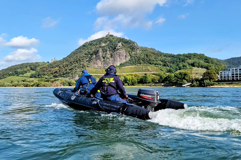 Ein kleines Motorboot fährt auf einem Fluss, darauf sitzen zwei THW-Kräfte in blauer Kleidung. Im Hintergrund befindet sich eine grüne Landschaft mit Bergen und Weinanbau.