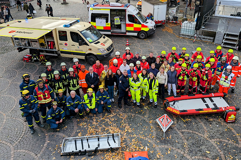 Ein Gruppenbild mit vielen Menschen in verschiedenen Einsatzkleidungen. Hinter der Gruppe stehen Einsatzfahrzeuge der Malteser und des Arbeiter-Samariter-Bunds. 