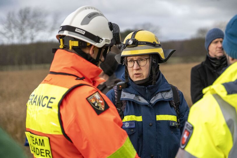 Mehrere Menschen stehen auf einem Feld. Zwei von ihnen tragen Schutzhelme. Auf dem Rückenschild des einen steht "France, Team Leader". (Quelle: THW/Robin Ramos-Hoffmann) Mehrere Menschen stehen auf einem Feld. Zwei von ihnen tragen Schutzhelme. Auf dem Rückenschild des einen steht "France, Team Leader".